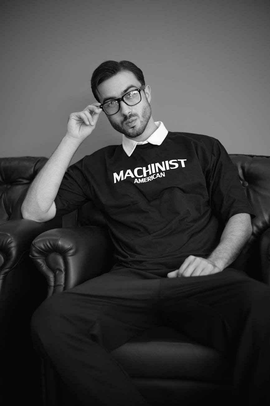 Man wearing eyeglasses and a Machinist American t-shirt, sitting on a leather armchair in a monochrome setting.